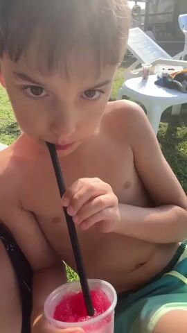 This is a close-up, eye-level shot of a young boy, likely of Middle Eastern descent, with brown hair and large, expressive brown eyes. He is shirtless, wearing green and blue swim trunks. The boy is holding a clear plastic cup filled with a pink, frozen beverage, and he is drinking it through a black straw. His left hand is holding the cup, while his right hand is also near it. He appears to be outdoors, with hints of a green lawn and lounge chairs in the blurred background. The lighting suggests it is daytime, with sunlight casting a glow on the boy's face and body. The overall mood is relaxed and summery, possibly indicating a day at a pool or beach. There is no visible text in the image.