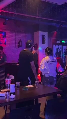 Several people are standing and conversing inside a dimly lit bar or club-like establishment. The lighting is colorful, with reds, purples, and greens illuminating the scene. A table with several plastic cups, bottles, and what appears to be an ashtray is in the foreground. Some people are gathered around this table. In the background, there’s a small stage with a lighted backdrop featuring a mural-style painting of four people. There are additional tables and chairs around the stage. People are talking to each other, and the atmosphere seems relaxed and social. The overall impression is of a casual gathering in an entertainment venue.