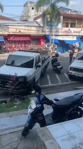 The video captures a street scene in Sumedang, Indonesia, likely during the daytime, judging by the bright sunlight. The setting is a commercial street lined with buildings. A prominent "HONDA" sign is visible on one of the storefronts. Another shop has a sign that reads "PO SUMBER HIDUP ELEKTRONIK" and appears to be an electronics store.

The street is populated with various vehicles, including several motorcycles and two white vehicles. In the foreground, a blue scooter is parked on a sidewalk, with its license plate visible. Behind it, a white pickup truck is parked, and further down the street, a white van or SUV is also present. A dark-colored motorcycle is parked between the pickup truck and the van.

There are at least two individuals visible. One person, wearing a dark blue t-shirt and black pants, is walking in the foreground, appearing to approach a motorcycle. Another person, wearing a dark shirt and lighter pants, stands near the white van.

The overall impression is of a typical urban street environment in Indonesia, with daily activities unfolding. The presence of businesses, vehicles, and people suggests a functional commercial area. The weather appears to be clear and sunny.