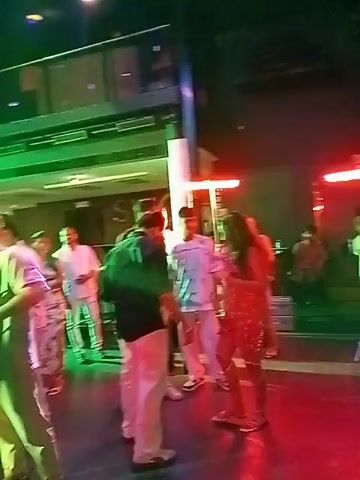 The video shows a crowded nightclub environment with several people mingling and standing around. The lighting is primarily blue, purple, and red, creating a vibrant, party atmosphere. The camera pans upwards to showcase the club's ceiling, decorated with a modern, chandelier-like structure featuring numerous strings of light. At the far end of the club, a large screen displays colorful, geometric patterns. A DJ booth is visible in front of the screen, suggesting live music is playing.