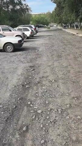 The video shows a dirt and gravel parking lot with several cars parked on the left side. The cars are facing away from the camera. The lot is lined with trees on the right side, and there appears to be a building in the distance. The ground is uneven and covered with loose rocks and dirt.