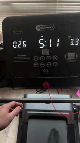 The video shows the console of a Dynamix treadmill in operation. The display reads a distance of 0.26, time elapsed is 15:81, and the speed is 3.3. Buttons for speed, program, mode, and numerical input are visible, as well as a start/stop button and a safety key with a red cord. The treadmill is running on a wooden floor, with blinds visible in the background. There is no person visible in the video.