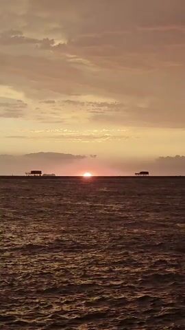 The video shows a calm sea with a sunset. Two small structures, possibly huts or piers, are visible on the horizon. The sky is filled with clouds, tinted orange and pink by the setting sun. The sun itself is a small, bright orb just touching the horizon. The water is dark and slightly rippled, reflecting the colors of the sky.
