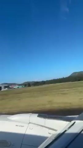 The video, shot from inside a plane, shows the aircraft landing. The plane wing, marked "NO STEP," is visible in the foreground. Outside the window, there's a clear blue sky above an airport runway surrounded by green fields and buildings. Mountains are visible in the background. The plane descends, approaching the runway.