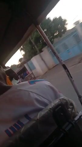 The video shows a first-person perspective from inside a motorized tricycle, also known as a "keke." The driver is wearing a grey and white shirt. The view is of the street outside the moving vehicle, including buildings, parked cars, and pedestrians.