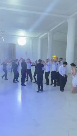 A group of men, dressed in formal attire including white shirts and black pants, are dancing in a large, well-lit room with a high ceiling and decorative columns. Some men are wearing black shirts and black pants. They are arranged in a circular formation on a polished floor that reflects the lights. The lighting in the room is a mix of white, blue, and yellow, with spotlights creating bright illuminated areas. Some of the men are raising their arms in the air and appear to be engaged in a lively dance, possibly a traditional group dance. There are tables and chairs visible in the background, suggesting that this event is a celebration or party.