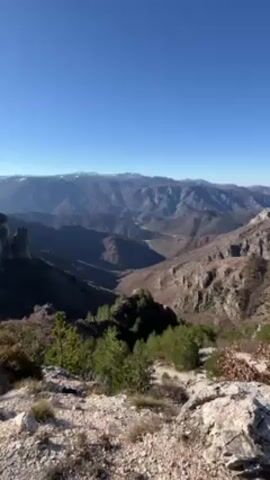 The video shows a mountainous landscape under a clear blue sky. The mountains are layered and stretch into the distance, with some snow visible on the peaks of the more distant ranges. The foreground features rocky terrain and sparse vegetation, including small trees and shrubs. A deep valley is visible between the mountains, with hints of a river or road winding through it. The scene is bathed in sunlight, with varying shades of shadow and light across the landscape.