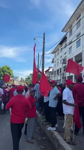 The video shows a street level view of a group of people gathered together, seemingly in a procession or rally. The majority of the people are wearing red shirts, and many are holding or standing near red flags. The flags vary in size and shape, some are small handheld flags, while others are large banners attached to poles. The crowd appears to be primarily men, although there are a few women visible. The people are of varying ages and skin tones. They stand in a line along the side of a street, and some people are chatting among themselves. Behind the crowd is a light-colored building, possibly a multi-story apartment or office building. The sky is blue with some scattered clouds. Power lines and streetlights are visible above the crowd. The street appears to be in a somewhat urban or suburban setting, possibly in a tropical location given the foliage.