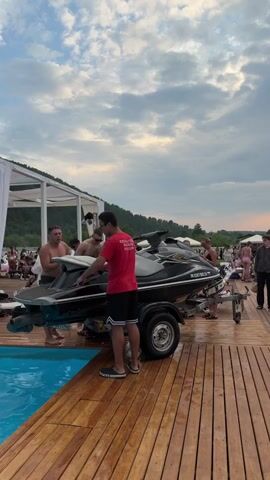 The media file captures an outdoor scene at what appears to be a waterside event or resort near Дрокино, Russia. The primary focus is a group of people gathered around a jet ski on a wooden dock.

In the foreground, a man in a red t-shirt and black shorts stands facing away from the camera, holding a red cup. To his left, two shirtless men are partially visible, one of whom appears to be talking and gesturing. Behind them, a jet ski on a trailer is the central object. Another person in a red t-shirt and shorts stands near the jet ski.

Further back, other individuals are present, some of whom are dressed in swimwear, suggesting a recreational or party atmosphere. A woman in a white dress and a cow-print jacket, wearing a cowboy hat, is prominently featured in the mid-ground, holding an orange cup and looking towards the camera. She is adorned with several bracelets and necklaces.

The setting includes a swimming pool to the left of the dock. In the background, there are tents or canopies, more people lounging, and a natural landscape with trees and hills. The sky is partly cloudy, indicating it is likely late afternoon or early evening given the soft lighting. The overall mood seems relaxed and social, typical of a summer gathering.

Visible text on the jet ski reads "PK 0267 BK 24," which appears to be a registration number. The red t-shirt worn by one of the individuals has text on the back, but it is too blurred to be legible.