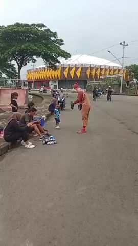 The video shows a clown interacting with a small child on a street. The clown is wearing an orange jumpsuit, red shoes, and a red hat, and has a painted face. The child is wearing a blue patterned shirt and shorts. The clown holds out a black bucket to the child, who seems hesitant. Several people, including children and adults, are sitting on a curb nearby, some of them looking at their phones. Behind them is a large tree and a skate park. In the background, there is a large building with a distinctive white and yellow facade. Traffic, including motorcycles and a bicycle, passes on the street. The weather appears overcast.