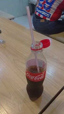 A partially empty bottle of Coca-Cola with a red cap and a pink straw sticking out of it is lying on a wooden surface. A person wearing blue jeans and a backpack with the words "SuperGros" is partially visible in the background.