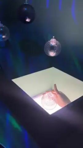 A person's hand is inside of a white square lit from below, holding a clear spherical ornament with ridges. Above the square are four similarly-shaped clear ornaments hanging from the ceiling. The wall behind the ornaments has a pattern of blue and green lights projected onto it.