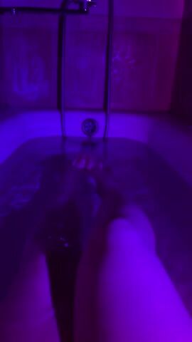 This is a first-person perspective video of someone in a bathtub filled with water. The scene is illuminated by purple lighting, creating a moody and atmospheric setting, likely a bathroom. The water is disturbed, indicating movement within the tub, and the person's legs are visible submerged in the water. The focus is on the legs and the water, with the drain and the showerhead visible in the background. The lighting suggests it might be nighttime or an indoor setting where the lights are deliberately set to this color. The overall impression is one of relaxation or self-care. There is no visible text in the media.