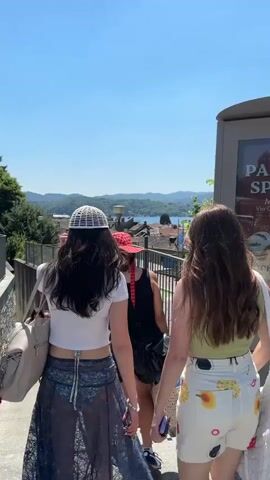 Two women walk up a set of outdoor stairs on a sunny day, away from the camera. The woman on the left has long, dark hair and is wearing a white crocheted cap, a white crop top, and a long, sheer skirt over what appears to be a swimsuit. She carries a beige tote bag on her shoulder. The woman on the right has long, brown hair and is wearing a green tank top and white shorts with colorful floral designs. In the background, a scenic view of a town with red-tiled roofs, a lake, and tree-covered hills can be seen. Several other people are visible on the stairs behind the women. The environment is bright and sunny with a clear blue sky. A railing and the side of a building are also visible.