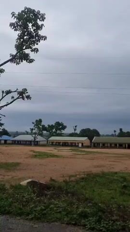 The video shows a landscape with buildings, trees, and a power line tower under a cloudy sky. The camera is moving, suggesting it is from a vehicle. The buildings appear to be simple structures, possibly a school or administrative complex, with visible roofs and walls. There are no people or animals visible in the frame. The environment is rural or semi-rural, with dirt paths and vegetation. The lighting is dim, indicating overcast weather.