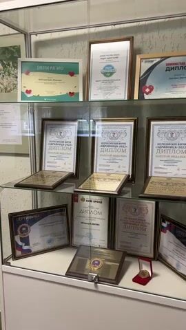 The video shows a glass cabinet displaying various diplomas, certificates, and awards. The cabinet has three shelves with a white frame. The certificates and diplomas are framed, mostly in gold or dark wood. Many of them are written in Russian. Some of the awards are medals displayed in presentation boxes. The camera pans slowly from left to right, showing each shelf of the cabinet.