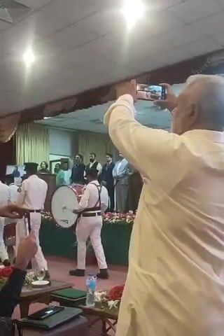 The scene captures an indoor event, likely a ceremony or presentation, with attendees seated and standing. A small band, dressed in white uniforms, parades into the room. A drummer is prominently featured. The room is lit with bright overhead lights and has an elevated stage area decorated with floral arrangements. Several individuals stand on the stage, observing the band's entrance. Many people in the audience record the event with phones and cameras. The room appears to be a large hall or auditorium.