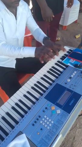 A man in sunglasses and a white shirt plays a Yamaha keyboard outdoors. The keyboard is on a metal stand. People stand and sit nearby, and there are tables with white tablecloths in the background. The ground is dirt. The man's hands move quickly across the keys as he plays. A phone, keys, and other items are on the keyboard.