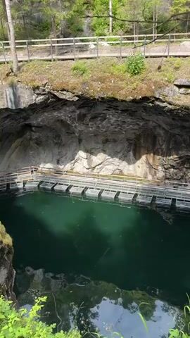 The video pans over a large, open-air quarry filled with dark green water. A wooden walkway with a railing is visible on the upper edge of the quarry, overlooking the water. Another walkway, made of metal grating and railings, extends over the water's surface, creating a bridge-like structure. The quarry walls are rough and rocky, with some vegetation growing on the edges. The overall impression is of a natural or man-made geological formation that has been made accessible to visitors. The lighting suggests it is daytime, with sunlight illuminating parts of the quarry walls. There are no people visible in the video.
