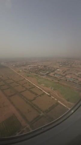 The video begins with an aerial view of a landscape from an airplane window. The ground below is a mix of agricultural fields and urban development, with a hazy sky. The camera then moves inside the airplane cabin, showing blurry images of passengers. The focus shifts to two young men sitting next to each other, both wearing neck pillows. One man is looking towards the camera and touching his face, while the other is also looking towards the camera with his hand raised. The video ends with a close-up of the airplane window, with bright light coming from outside.