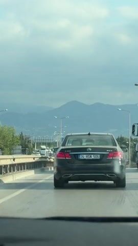 The video shows a rear view of a black Mercedes-Benz sedan driving on a highway. The car has a license plate that reads 34 HSN 506. In front of the Mercedes, there is heavy traffic with other cars and a white tanker truck to the right. In the background, there are mountains and a cloudy sky. The road appears to be wet.