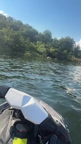 A first-person perspective video shot from a watercraft shows a body of water with ripples. The watercraft has a dark and white exterior. In the distance, a lush green shoreline is visible with trees and a hilly landscape. Further out on the water, another boat is seen from the stern. The sky is blue with scattered white clouds. The video shows the watercraft moving through the water, with the shoreline and the other boat remaining in view.
