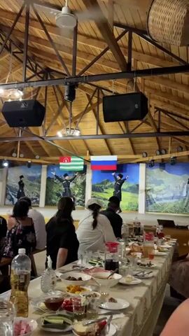 The video shows a group of people seated at a long table laden with food and drinks, watching a performance on a stage. The stage features dancers in traditional attire, possibly performing a folk dance, in front of a backdrop depicting mountainous landscapes. Two flags, one appearing to be the flag of Abkhazia and the other the flag of Russia, are displayed prominently on the stage. The setting appears to be an indoor venue with a wooden ceiling and exposed beams, and speakers are mounted overhead. The camera is positioned from the perspective of someone seated at the table, looking towards the stage.