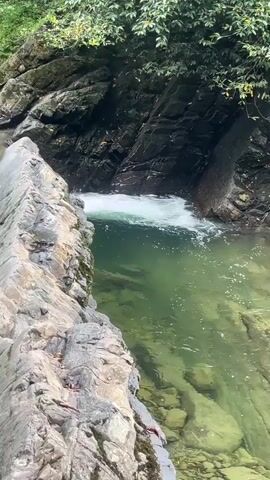 A narrow, rocky gorge with a pool of clear, green water at the bottom. A small waterfall spills into the pool from a crevice in the rock face. The water is turbulent where the waterfall enters, with white foam visible. Lush green foliage overhangs the gorge from above. The image is shot from a slightly elevated perspective, looking down into the gorge. The lighting suggests it is daytime, with shadows indicating the presence of trees or other obstructions blocking direct sunlight.
