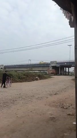 The video shows a person on a bicycle on a dirt road with a bridge in the background. The person is wearing dark clothing and is riding the bicycle away from the camera. The bridge is elevated and appears to be made of concrete. There are power lines strung across the sky above the bridge. The area around the road is overgrown with vegetation, and there is some trash scattered on the side of the road. The sky is overcast, suggesting a cloudy day. The video is shot from a slightly elevated perspective, looking down on the scene.