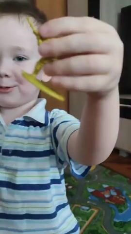A young boy with short brown hair and blue eyes, wearing a white and blue striped polo shirt, is holding a Batman figure in his left hand and a small, yellow, stick-like object in his right hand. He is smiling at the camera. The Batman figure is black and gray with a yellow utility belt and a black cape. The boy appears to be indoors, with a colorful play area visible in the background. A closet with mirrored doors is visible behind him.