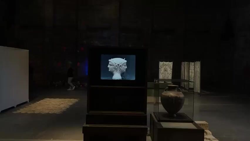 The video depicts a dimly lit exhibition space, likely an art gallery or museum in Venice, Italy. In the center, a monitor displays a static image of a classical-style, white, double-faced bust, resembling Janus, with two faces looking in opposite directions. The bust has a weathered texture and distinct classical features, including horned elements on its head.

To the right of the central display, an antique-looking, dark brown ceramic urn or vase with handles is showcased within a glass display case, resting on a wooden base. Further back on the right, two panels of patterned fabric or tapestries hang, adding to the decorative elements of the exhibit.

On the left side of the frame, a white panel or wall is visible. A person is seen walking in the background, partially obscured and moving from left to right, their presence indicated by subtle light reflecting off them. The floor is dark, and in the mid-ground on the left, a patch of lighter-colored, possibly textured material, is laid out. The overall lighting is low, with strategic illumination highlighting the exhibits. The background also features faint, shifting colored lights, including blue, green, and red, creating a dynamic, atmospheric effect against the dark walls.