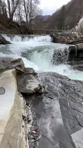 The video depicts a rocky stream with a series of small waterfalls. The water is white and foamy as it rushes over the rocks.  Bare trees line the banks of the stream, and a bridge is visible in the background. The sky is partly cloudy. The camera moves slowly, capturing the flow of the water and the rugged landscape.