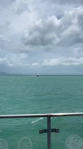 The video shows a ferry boat moving across a choppy, turquoise-colored sea under a cloudy sky. The perspective is from a lower deck of another boat, indicated by the presence of a metal railing in the foreground. The ferry is the primary object of focus, positioned in the center of the frame as it cuts through the water, leaving a white wake behind it. A distant landmass is visible on the horizon beneath the clouds.