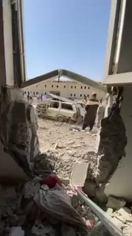 The video reveals a scene of significant destruction, apparently caused by an explosion. The shot is taken from inside a building, looking out through a large hole in the wall, likely where a window once was. Debris, including chunks of concrete, blocks, and twisted metal, litters the ground. In the center of the scene, a badly damaged white station wagon can be seen, with parts of its body missing or severely deformed. In the background, a building with many windows is visible, suggesting it could be an office or residential structure. Several people are present, some dressed in traditional Muslim attire, moving around and examining the damage. The sky is clear and blue, indicating a daytime setting.