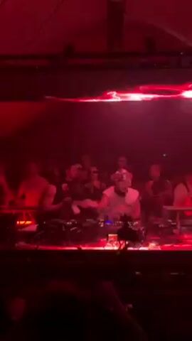 The video shows a DJ performing at a club. They are wearing a white shirt, glasses and headphones and standing behind a console with a lot of equipment and cords. Several people are standing behind the DJ. The lighting is red and there is a large screen above the DJ, also lit up red. The overall setting appears to be a dark club with a lively atmosphere.