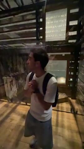 The video shows a person standing in a room with rough-hewn wooden walls and ceiling. The person is wearing a white t-shirt, dark shorts, and a black backpack. They are holding something in their hands and looking upwards towards a display on the wall. The display appears to be a screen with text on it. The room is dimly lit, with spotlights illuminating the display and the wooden interior. There are other displays visible on the wall to the left of the person, featuring framed images or documents. The floor is made of wooden planks. The overall impression is of a museum exhibit or historical reconstruction.