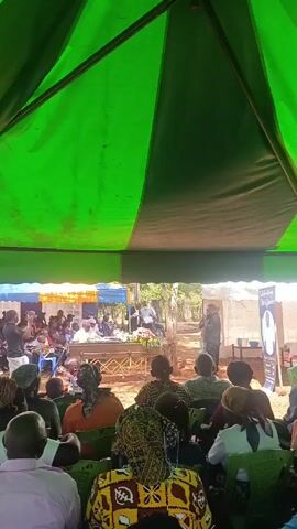 The video depicts an outdoor gathering, presumably a funeral or memorial service, taking place under a large tent with a green and black striped canopy. The camera is positioned from the back of the crowd, providing a view of the attendees seated on green plastic chairs. A man is speaking into a microphone, addressing the crowd gathered around a casket decorated with flowers. In the background, beyond the gathered attendees, there are trees and a small building. A banner stands beside the speaker, featuring a portrait and text that reads "Celebrating Life." The overall setting suggests a somber and respectful atmosphere.