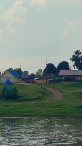 This video shows a view of a rural village from across a body of water. In the foreground, there is a body of water with ripples on its surface. The middle ground features a grassy embankment with a dirt path winding through it. Several houses and buildings are visible on the embankment, some with blue roofs. There is also a small gazebo-like structure with a blue roof. In the background, a tall cell tower stands prominently, and a larger wooden building with many windows is visible. Trees and vegetation are scattered throughout the scene. The sky is partly cloudy with a blue sky visible. The overall impression is of a tranquil, rural landscape.