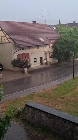 The video displays a residential street scene in Klettgau, Germany, during a heavy rain shower. The focus is on a two-story house with a terracotta-tiled roof and light-colored walls, featuring a prominent gable with exposed timber beams on the left side. A large garage door with pink accents is visible, adjacent to a rose bush in bloom. A small entryway with steps leads to the main door of the house, and a large, leafy tree stands to the right of the house, casting a shadow on the wet street.

The street itself is asphalt, glistening with rain, and a paved sidewalk runs along the edge. The weather is overcast, typical of a rainy day, and the rain is clearly visible as streaks falling from the sky, creating ripples on the wet surfaces. A traditional street lamp stands on the right side of the road. In the foreground, a stone wall with some greenery and a large rock adds a natural element. The overall mood is tranquil, perhaps even a bit melancholic, due to the heavy rain. No people are visible in the frame, and there is no visible text in the media. The time of day appears to be daytime, given the ambient light, but the overcast sky makes it difficult to determine precisely.