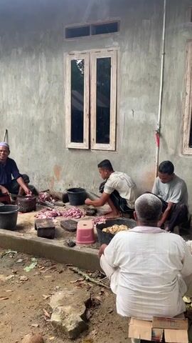 Four men are gathered outdoors near a concrete building. The building has a rough, grey textured wall and two double-paned windows. In the foreground, the ground is dirt and littered with leaves and debris. The men are engaged in preparing meat. One man on the far left, wearing a blue shirt and white head covering, is chopping meat with a large knife. In front of him are buckets and piles of cut meat. To his right, another man in a white t-shirt is also working with meat. Further to the right, a third man in a grey shirt is seated and appears to be supervising or preparing to help. In the immediate foreground, facing away from the camera, a fourth man in a white shirt is seated on a cardboard box, observing the activities. Large containers filled with meat and parts of an animal are visible. The overall setting appears to be a communal activity, likely related to food preparation.