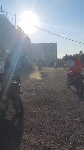 A sunny outdoor scene shows people on dirt bikes. In the foreground, a person in dark clothing on a dirt bike is partially visible, with their bike's front tire prominent. Behind them, another person in an orange shirt and black pants on a dirt bike turns their head to the left. Further in the background, a person in a red shirt on a dirt bike rides away from the camera, kicking up dust. More people and dirt bikes are visible in the distance, along with a large building and a ramp structure. The ground is covered in gravel. The sun is bright and high in the sky.