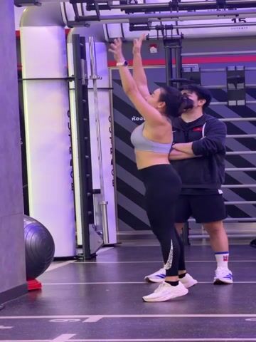 A woman in athletic wear is performing a wall ball exercise with a large black medicine ball in a gym. She is in a squatting position, preparing to throw the ball upward. A man stands behind her, observing. The gym environment includes fitness equipment and a wall with a mirrored surface.