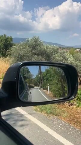 The video shows a view from inside a car, looking into the passenger side mirror. The mirror reflects a winding asphalt road bordered by dry grass and green foliage. The road curves to the left and leads towards a distant power line pole. The sky above is blue with fluffy white clouds. The side of the car and the driver's side window are partially visible in the foreground. The car appears to be in motion, as indicated by the blurred dry grass and the slight shift in perspective.