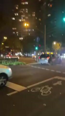The video shows a street scene at night. Traffic lights are green, and cars are crossing an intersection. There are tall buildings in the background, illuminated by lights. A designated bike lane is marked on the street. The video appears to have been taken from a sidewalk.