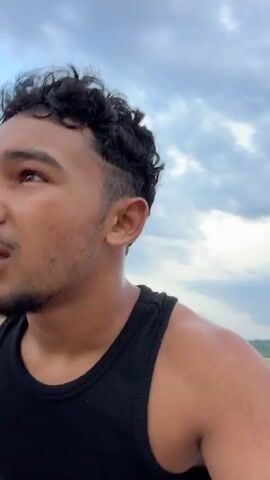 A close-up, low-angle shot shows a young man with dark, curly hair and a beard wearing a black tank top and olive green shorts. He is leaning forward, looking to his left, with his mouth slightly open as if speaking. The background is blurred, with hints of a cloudy sky and a dirt road or path. The man appears to be riding a vehicle, possibly a dirt bike, as a handlebar with a pink grip is visible in the foreground. The camera perspective suggests the footage is being captured from a moving vehicle. The man's facial expression and the motion blur indicate a dynamic and possibly fast-paced activity.