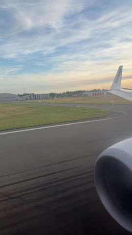 The video captures a view from an airplane window as it taxis on a runway. The plane's engine housing is visible in the foreground, and the wing with the Ryanair logo is on the right. The runway stretches out ahead, with tire marks from previous takeoffs and landings visible on the tarmac. Beyond the runway, there is a grassy field, followed by a row of hangars or airport buildings. In the distance, a control tower and other airport structures can be seen against a sky with scattered clouds and hints of sunset. The overall scene depicts the typical environment of an airport tarmac just before takeoff or after landing.