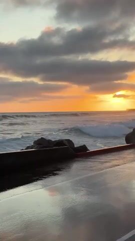 A video shows a sunset over the ocean. The sky is filled with orange and gray clouds. Waves crash against a rocky shore. A wet walkway runs along the edge of the water, reflecting the sky. Two figures walk along the walkway, silhouetted against the sunset.