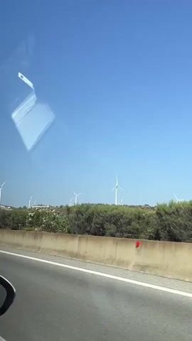 The video shows a view from a vehicle moving along a highway. In the foreground, a concrete barrier separates the highway from a grassy area. Beyond the barrier, a line of wind turbines is visible against a clear blue sky. The turbines are white with three blades each and are spread out across a gentle slope. Green bushes and some scattered white flowers grow between the turbines. The sun is shining brightly, casting shadows and creating reflections on the vehicle's window, which is visible in the upper left portion of the frame. A portion of the vehicle's side mirror is visible in the lower left corner.