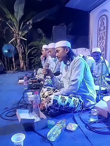 The video shows a group of men, likely religious scholars or performers, sitting on the floor in a dimly lit room. They are all wearing white caps and traditional clothing. Some of them are holding microphones and appear to be singing or chanting. There are also bottles of water, cups, and other items scattered around them on the floor. The lighting in the room changes colors, from blue to purple to red and green, creating a dynamic visual effect. In the background, there is a banner with text in Indonesian that appears to be related to a religious event. The overall atmosphere seems to be one of solemnity and cultural celebration.