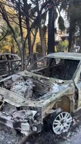 The video shows a scene of destruction following a fire. The primary focus is on the charred remains of what appears to be a car. The vehicle is heavily damaged, with the interior completely burnt out and the metal frame distorted and blackened. The tires and wheels are still visible, though also damaged. Other burnt vehicles are visible in the background. The surrounding environment shows signs of fire damage as well, with charred trees and debris scattered around. The sky is overcast, and the overall atmosphere is one of devastation.